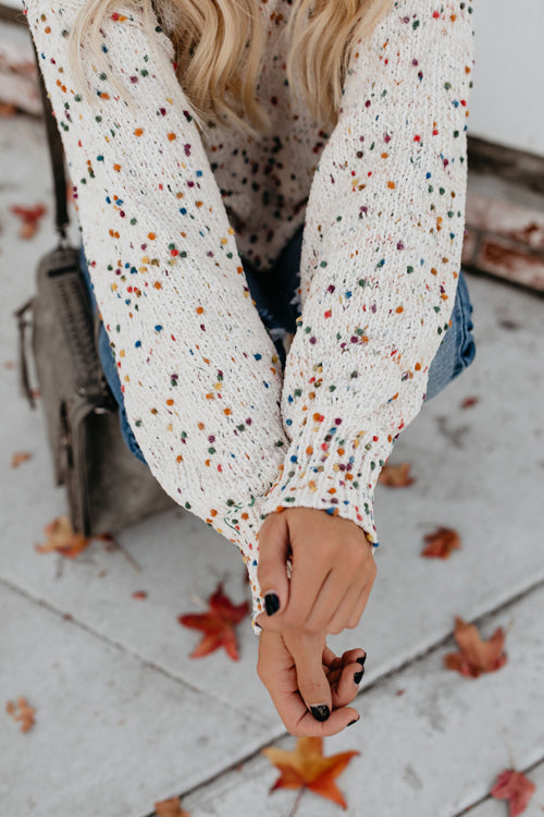 Beige speckled dot knit sweater sleeve and hem detail, hands at waist