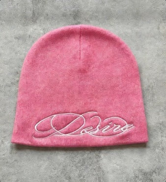 Pink knit beanie with white cursive “D” style letter embroidery, flat lay.