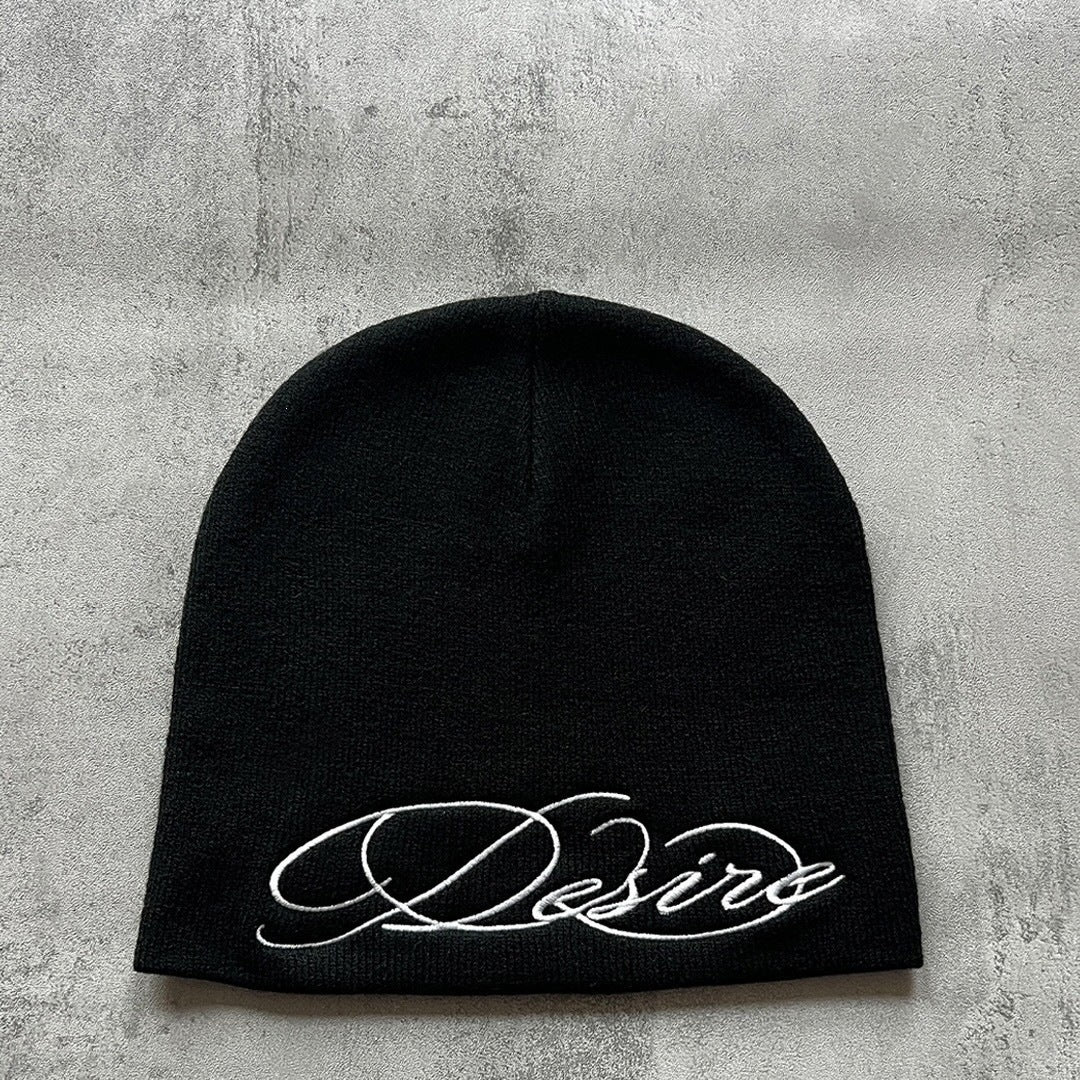 Black knit beanie with white cursive “D” style letter embroidery, flat lay.