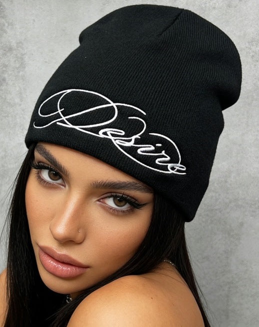Black slouch knit beanie with white cursive “D” style letter embroidery.